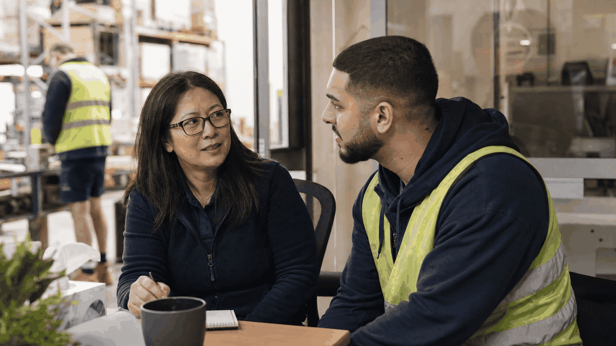 A manager sits and talks with a worker in a hi-vis vest at a table in a warehouse break area, taking notes.