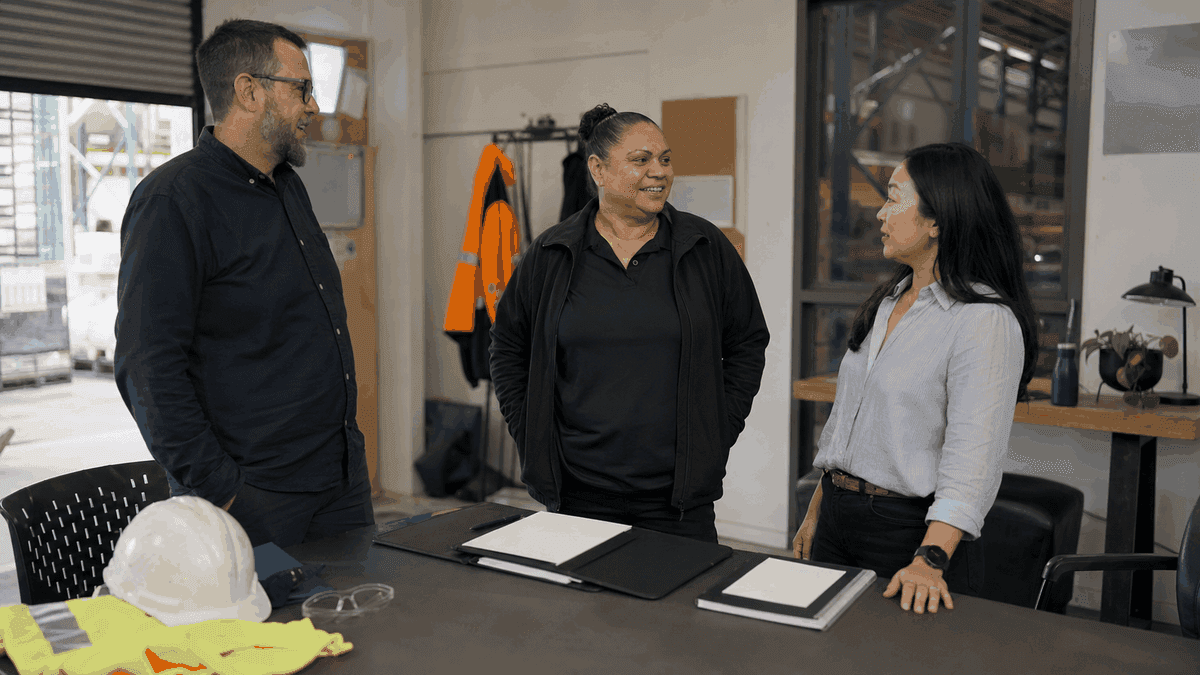 Three team members stand and talk in a light site office, with a hard hat, hi-vis vest and claim folders on the table.