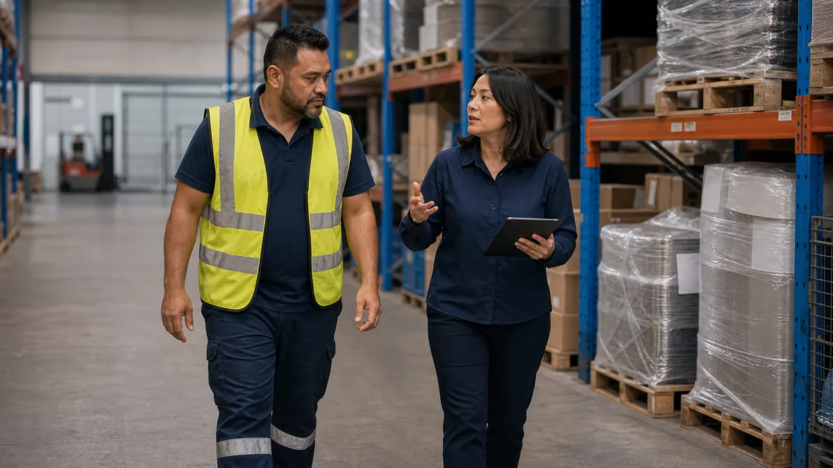 An injury management consultant walks a warehouse aisle with a worker in a hi-vis vest, talking as she holds a tablet.